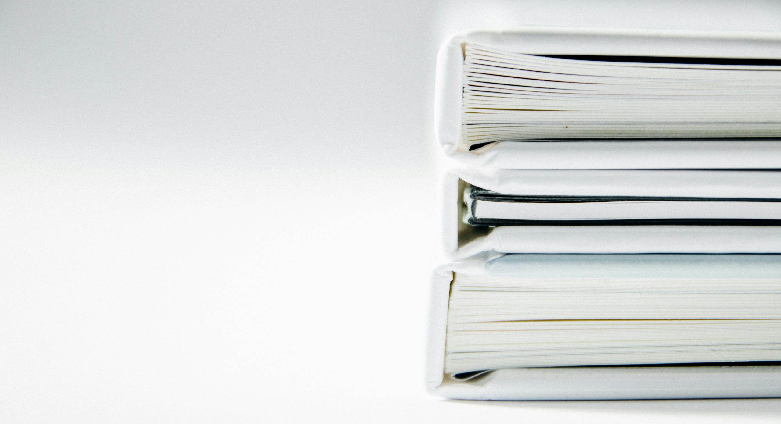 A partial view of a stack of 3 white binders filled with paper. White and grey background.