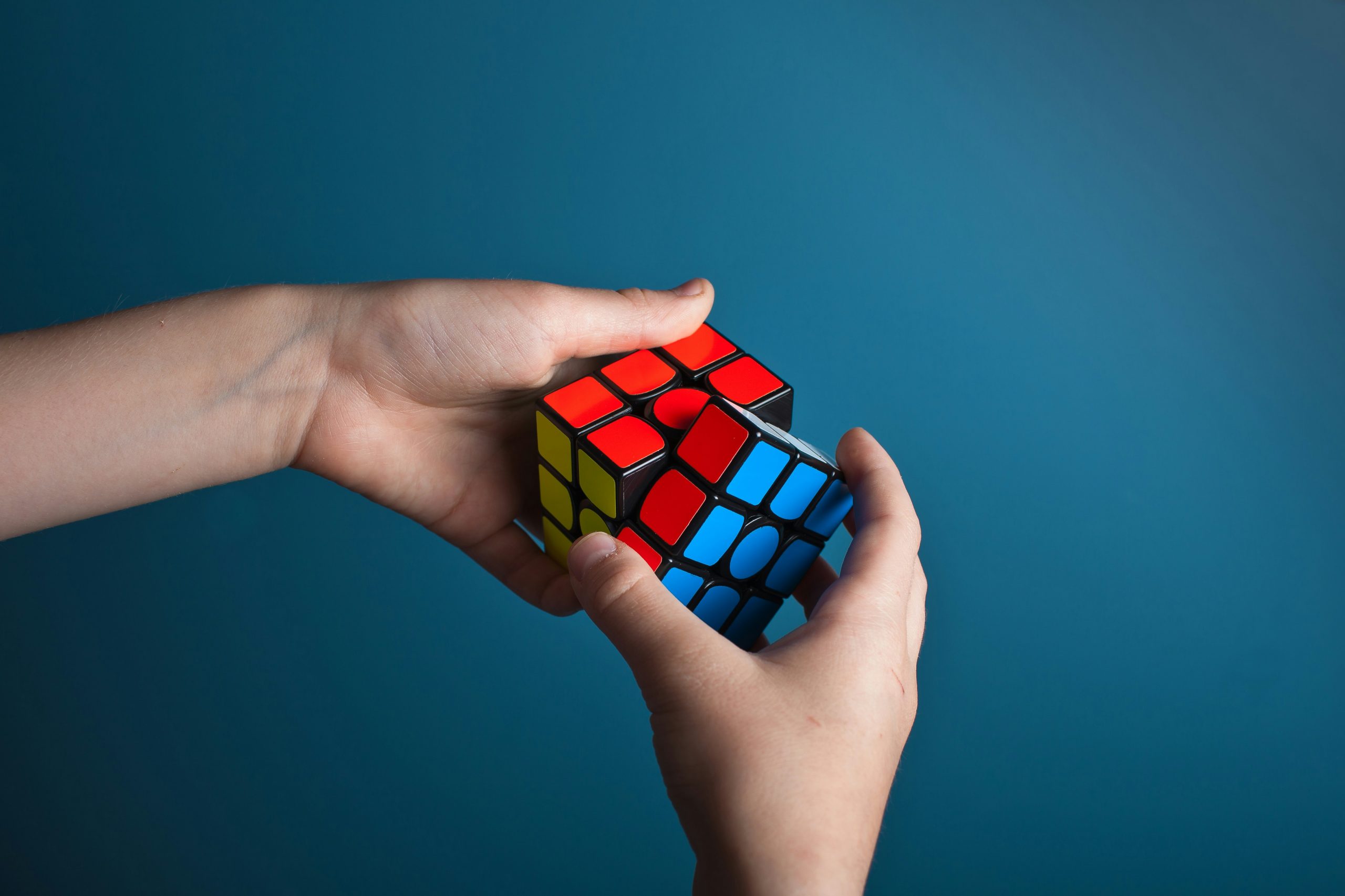 An almost-solved Rubix cube being turned by two hands. A blue background.
